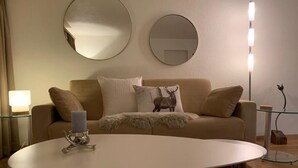 Olohuone, jossa on beige sohva, kaksi pyöreää peiliä, lasinen sohvapöytä ja lattiavalaisin.