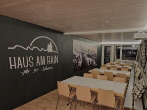 Un espace repas avec des tables et des chaises, un mur orné d’une fresque représentant un paysage de montagne, et un logo portant l’inscription « HAUS AM RAIN Alt St. Johann ».