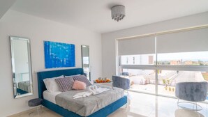 Une chambre moderne avec un grand lit, deux fauteuils, une tête de lit bleue, une grande fenêtre et une vue sur la ville.