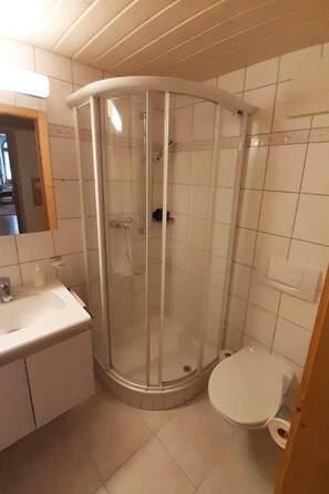 Une petite salle de bain située dans un coin, avec une douche à parois de verre, un lavabo blanc et des toilettes.