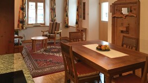 Ein Essbereich mit Holztisch, Stühlen und Teppich. Es gibt Fenster mit Vorhängen, eine Holztür mit Spiegel und einen kleinen Tisch mit Obst.