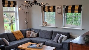 Een moderne woonkamer met een grijze modulaire bank, een houten salontafel en een zwarte leren fauteuil. Er zijn twee ramen met zwart-wit gestreepte gordijnen, en een wandlamp.