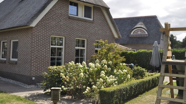 Een bakstenen huis met een steil rieten dak, een klein raam en een tuin met hortensiakriebels.