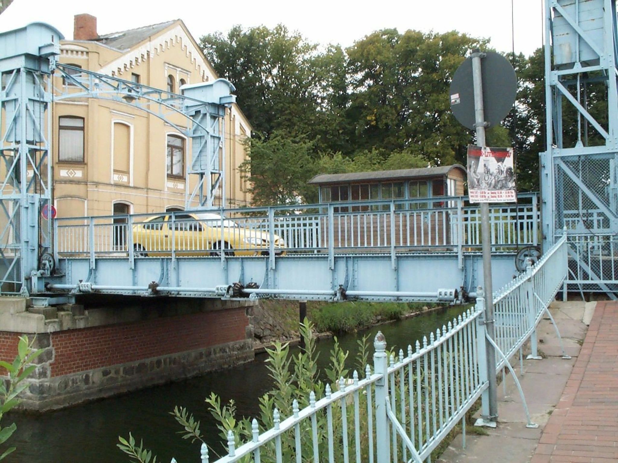 A bridge with a yellow car crossing over a canal, surrounded by greenery and buildings.