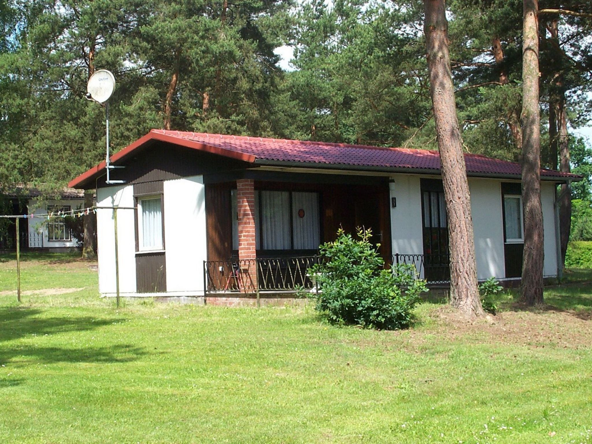 Ein eingeschossiges Haus mit rotem Dach, einem Backstein-Schornstein und einer kleinen Veranda mit schwarzem Geländer.
