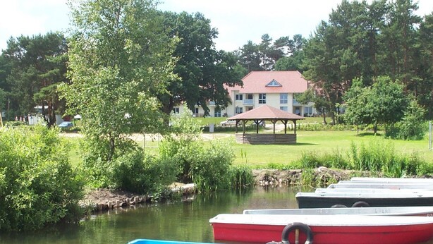 A row of boats docked by a calm river, a gazebo, and a building with a red roof in the background. - mit Garten, Terrasse und Grill by Interhome (Plau am See)