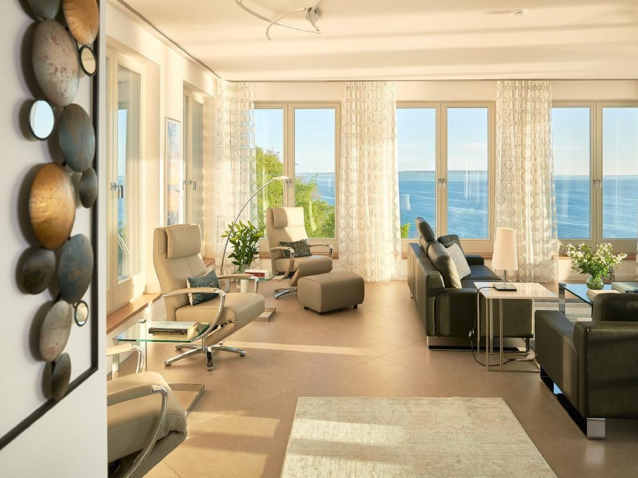 A modern living room with a view of the sea, featuring a sofa, armchairs, a coffee table, and a dining area.