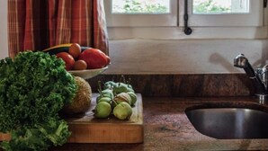 Una cocina con encimera de madera, un lavamanos, una ventana con cortinas y una variedad de verduras frescas.