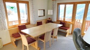 Une pièce chaleureuse avec une table et des chaises en bois, de grandes fenêtres avec des rideaux et une vue sur l’extérieur.