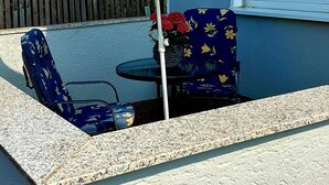 Um pátio externo com um guarda-sol floral azul, duas cadeiras azuis, uma mesa pequena e um vaso com planta.