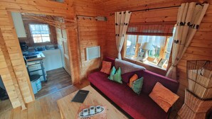 Hyggelig kabineinteriør med træpaneler, en sofa med puder, et spisebord, et køkken og et vindue med gardiner.