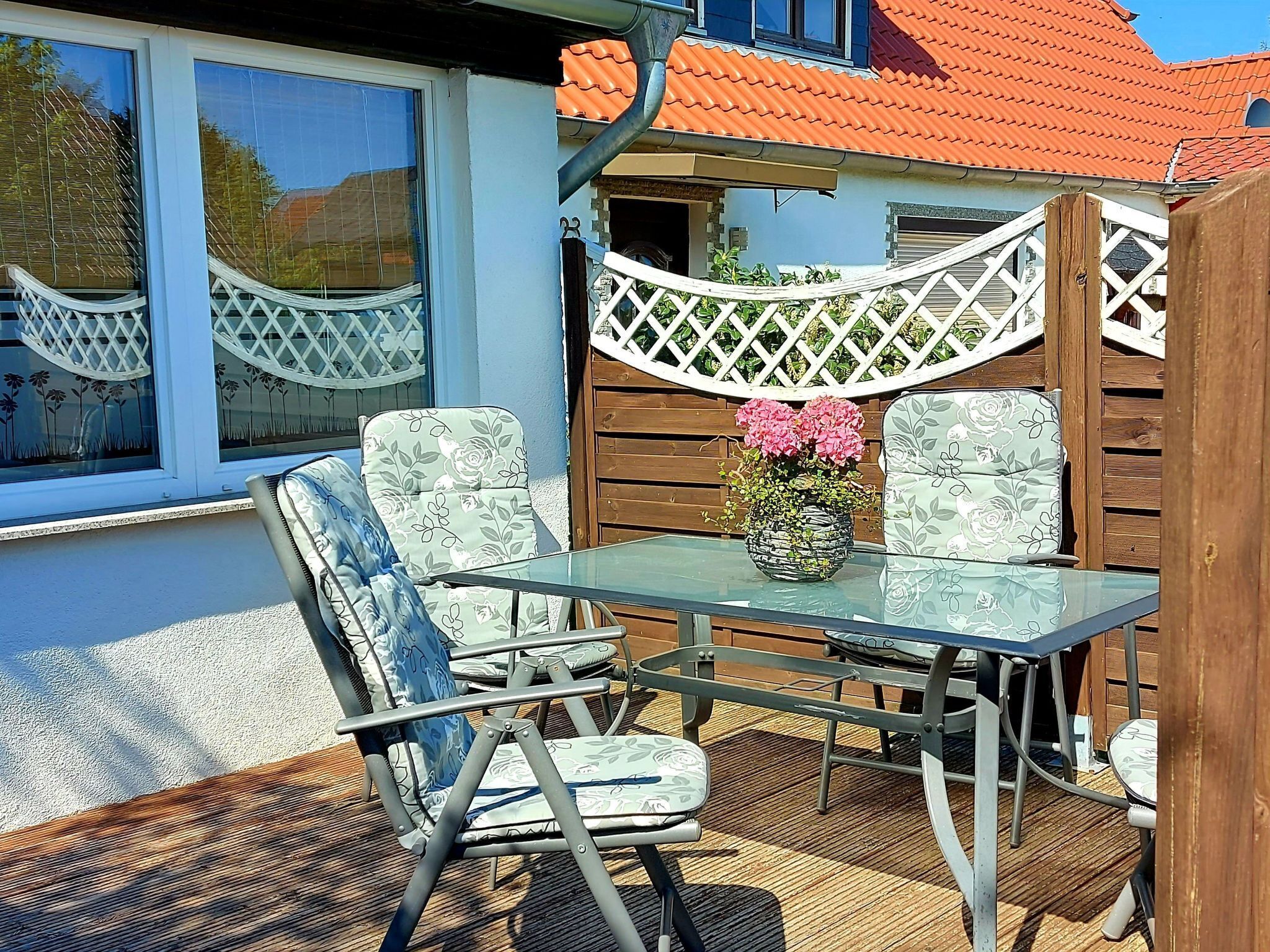 An outdoor patio with a glass table and four chairs, a wooden fence, and a window with a lattice design.