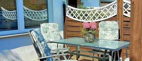 An outdoor patio with a glass table and four chairs, a wooden fence, and a window with a lattice design.