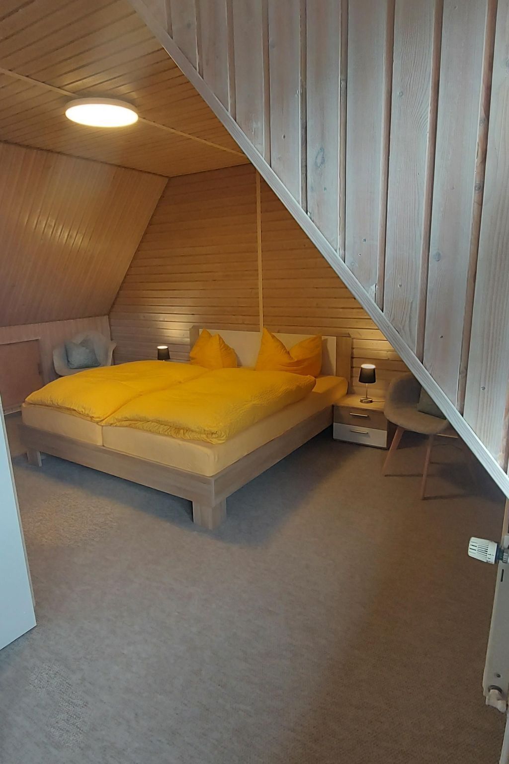 A bedroom with a sloped ceiling, a bed with yellow bedding, a bedside table with a lamp, and a chair.