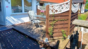 Une maison avec le numéro 23 sur la porte, une terrasse avec une table et des chaises, et une clôture en bois ornée de plantes grimpantes.