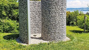 En cylindrisk stenhus med metalramme, omgivet af grøn vegetation og en klar himmel.