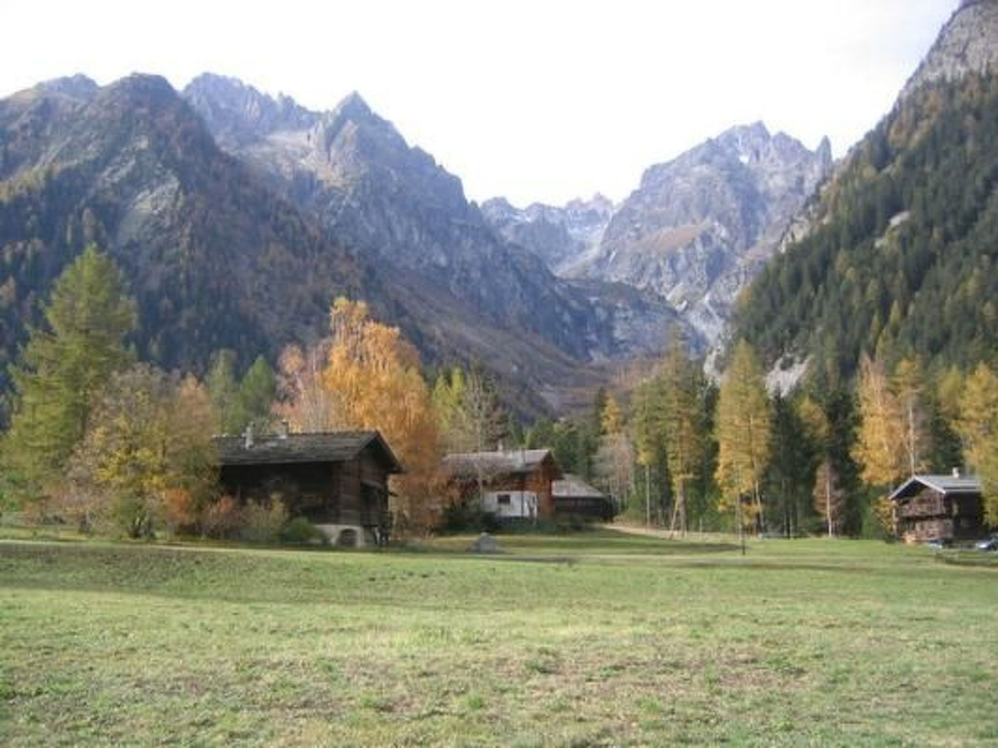 A mountainous landscape with two wooden cabins, one with a visible chimney, surrounded by trees and a grassy field.