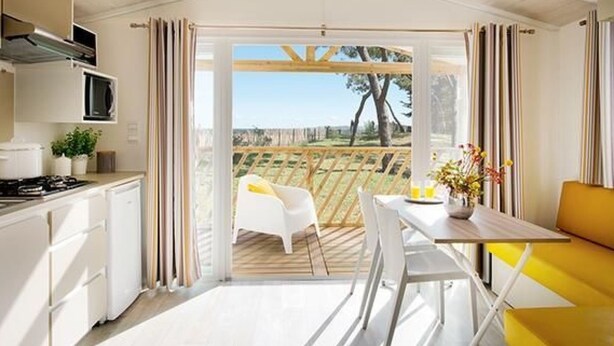 A modern kitchen with white cabinets, a stove, and a microwave. There is a dining area with a yellow chair and a table, and a view of a garden through large glass doors. - in Hohenlychen mit Grill, Loggia 3 by Interhome (Lychen)