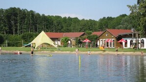 Un cadre idyllique au bord d’un lac, avec une tente, un petit bateau et des bâtiments aux toits rouges dotés de terrasses extérieures.