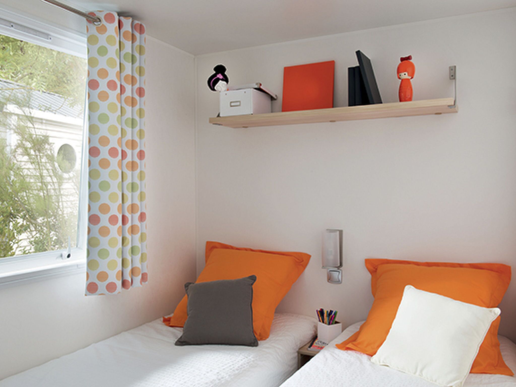 A bedroom with two beds, each with an orange pillow and a grey pillow, a window with polka dot curtains, and a shelf with various items. - Mobilheim Lagune II in Lychen by Interhome (Lychen)