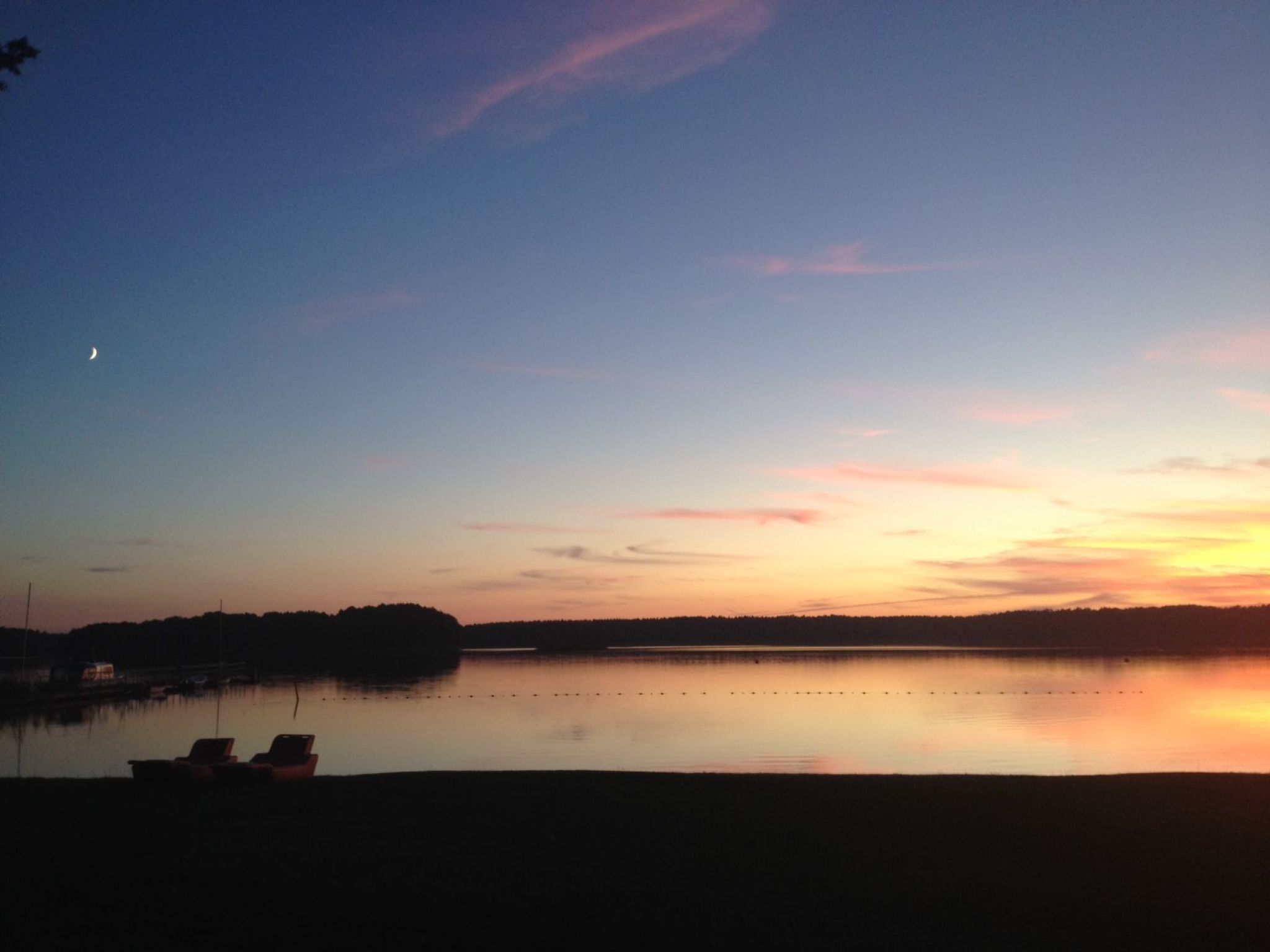 A calm lake at dusk with a crescent moon visible in the sky.