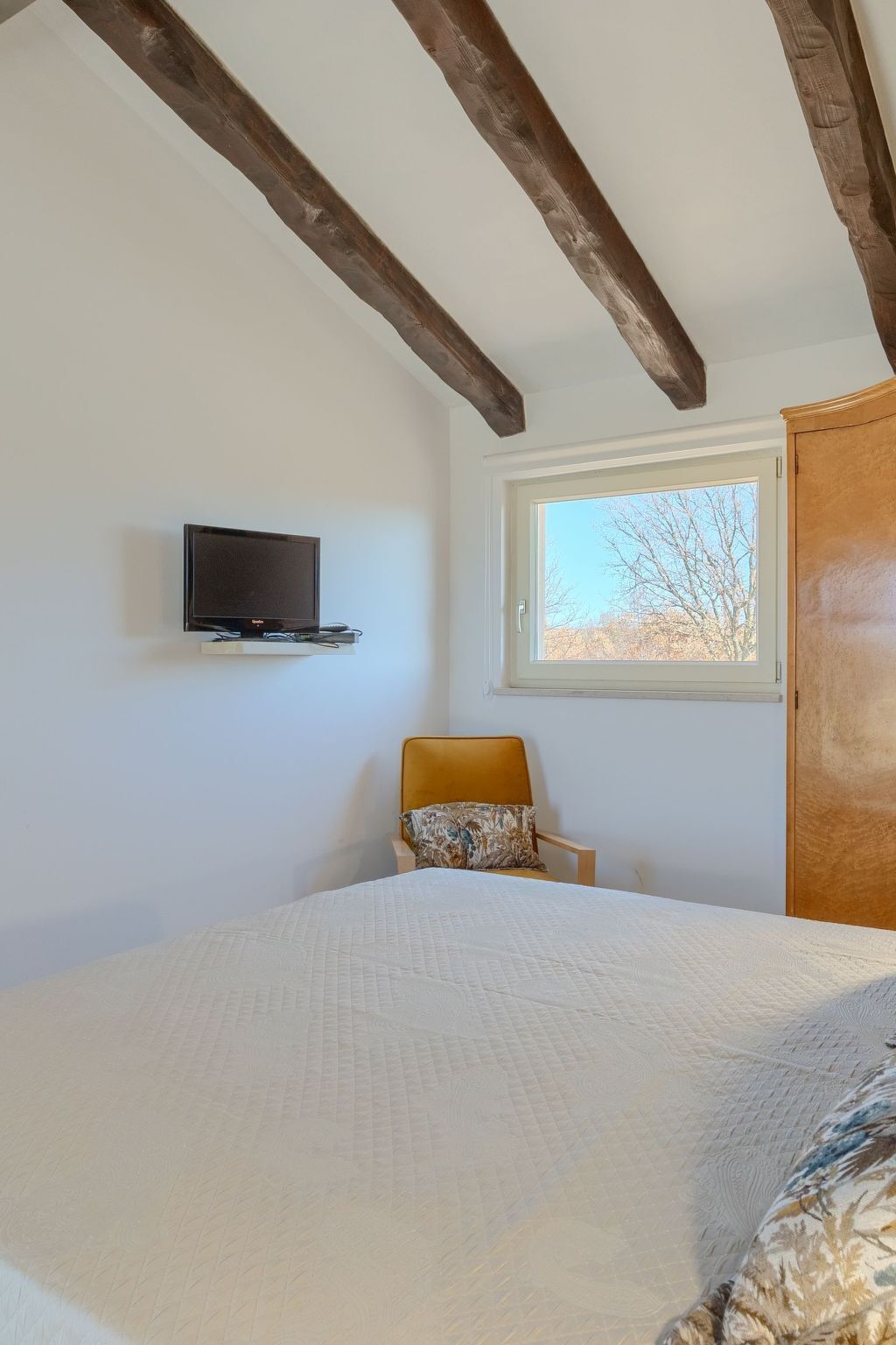 A bedroom with a bed, a chair, a television mounted on the wall, and a window with a view of trees.
