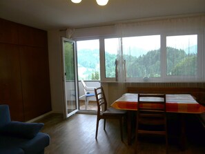 Une pièce comprenant une table à manger, des chaises, un canapé bleu et une vue sur une forêt, visible à travers de grandes fenêtres.