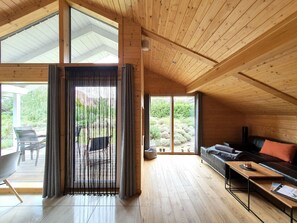 Quarto amplo com piso de madeira e teto inclinado, com sofá, mesa de jantar e uma grande janela com cortinas.