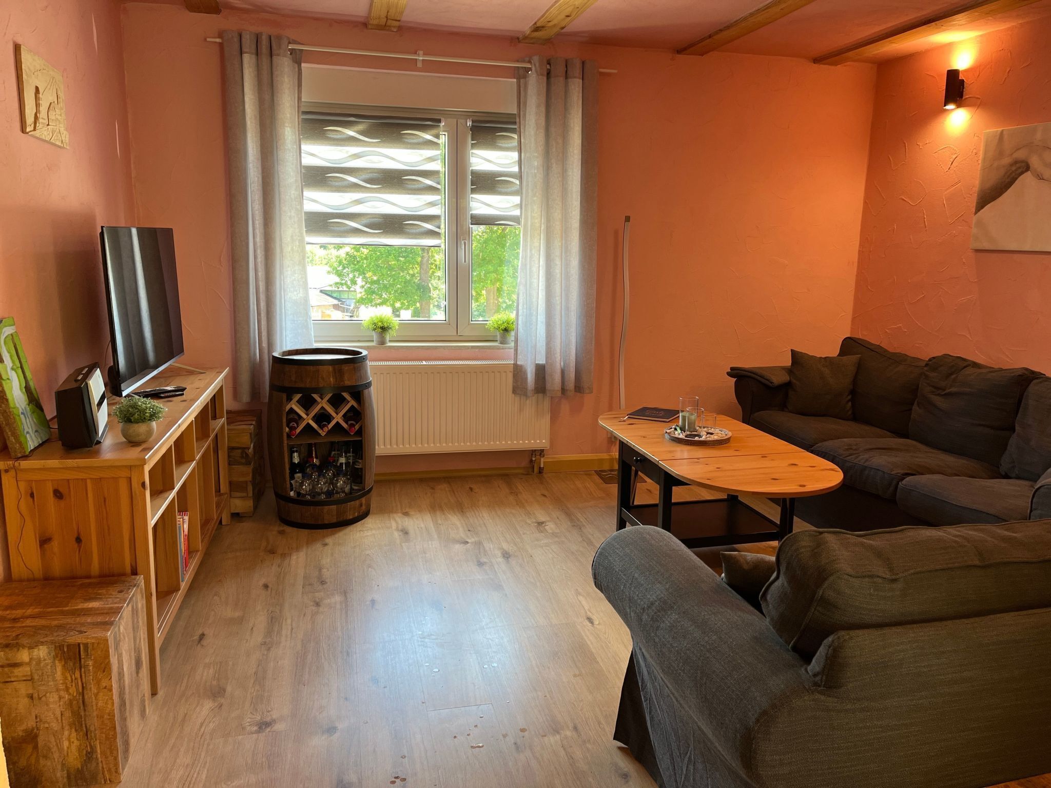 Un salon chaleureux avec un plancher en bois, un canapé, un téléviseur posé sur un meuble en bois, un tonneau à vin et une fenêtre avec des rideaux.