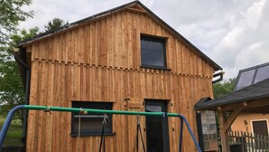 Et træhus med et stort vindue og en dør, et gyngestativ foran og et lille skur med et vindue.