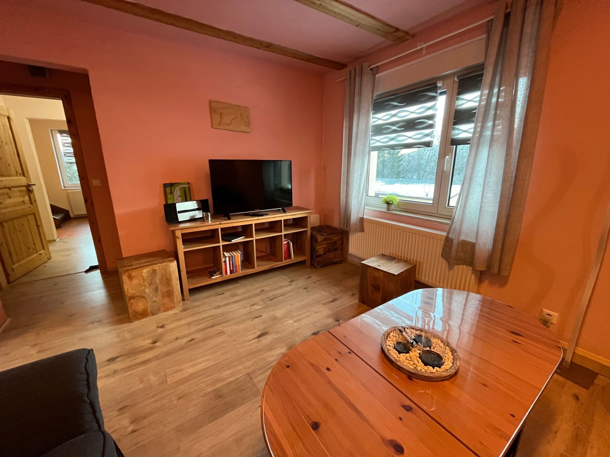 A cozy living room with a wooden TV stand, bookshelves, a wooden table, and a window with curtains.