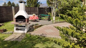 Un patio trasero con un horno de pizza a leña, una mesa con mantel floreado, un banco verde y una carretilla.