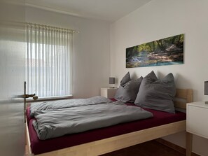 A bedroom with a bed, a window with sheer curtains, a painting of a forest scene, and bedside tables with lamps. - Hans am Klostersee by Interhome (Dargun)