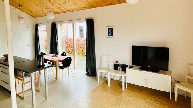 En rummelig opholdsstue med træloft, et spisebord med stole, en hvid sofa, et fjernsyn og et lille bord med en vase med blomster.