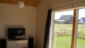 En stue med et fjernsyn på et hvidt skab, højtalere og udsigt til et hus gennem en skydedør.