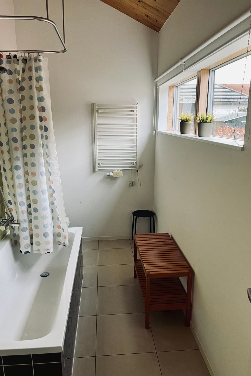 A small bathroom with a bathtub, a wooden stool, a radiator, and a window with potted plants.