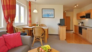 Een moderne woonkamer met een bank, een eettafel en een schaal fruit. In de achtergrond is een keuken met houten kasten en apparatuur zichtbaar.