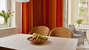 En matplats med ett bord dukat för två personer, en skål med frukt, flätade stolar, en vit hänglampa och utsikt över utomhusmiljön genom ett fönster med gardiner.
