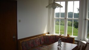 Een eethoek met een houten tafel en bank, een hanglamp en uitzicht op een groen landschap door grote ramen.