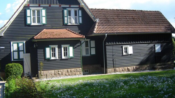 Een twee verdiepingen tellend huis met een steile kap, groene luiken en een stenen fundering.