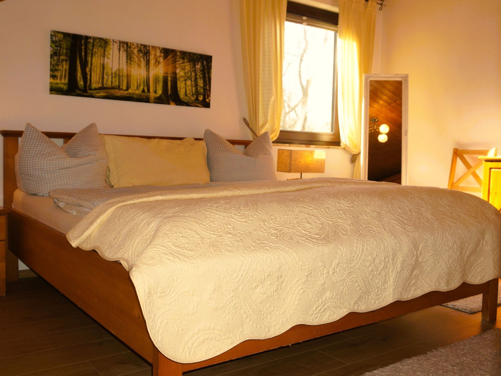 A bedroom with a wooden bed, a large window with curtains, a painting on the wall, and a bedside table with a lamp.