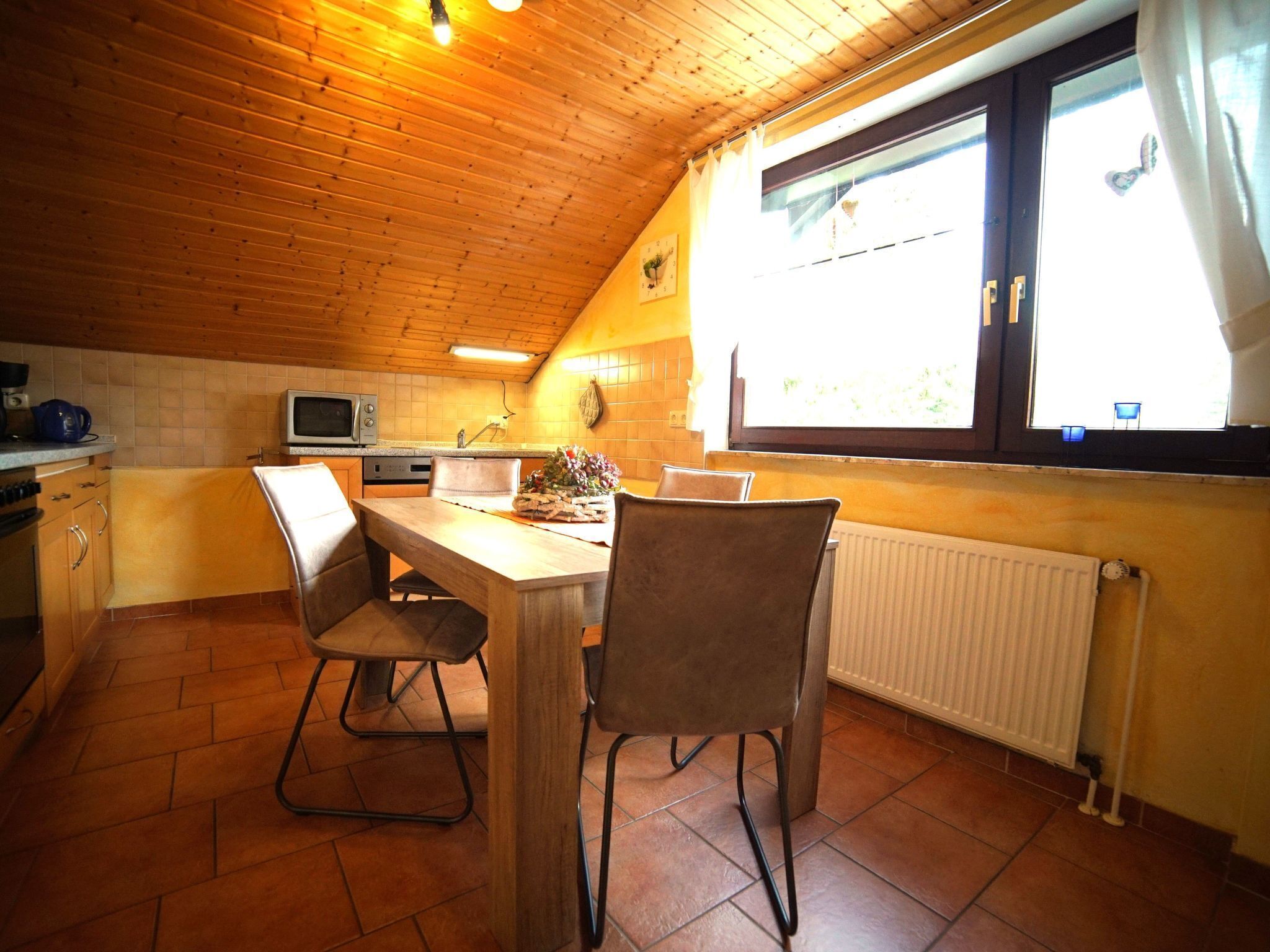 A cozy kitchen with wooden ceiling and walls, a dining table with chairs, a microwave, and a window with curtains.