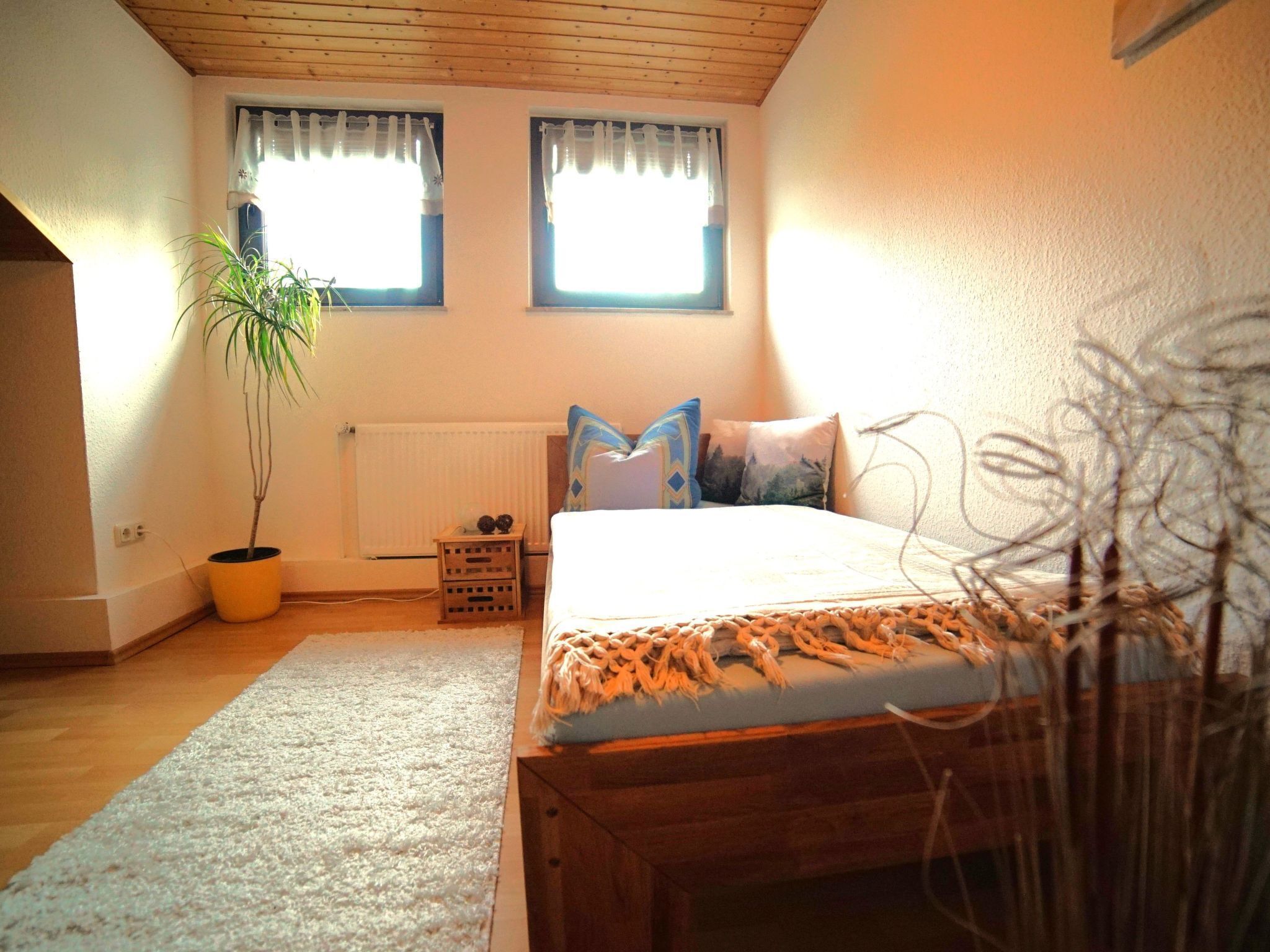A bedroom with a wooden ceiling, two windows with curtains, a bed with a patterned bedspread, a potted plant, and a small wooden nightstand.
