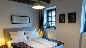 Ein ordentlich bezogenes Bett mit floralem Überwurf, ein Nachttisch mit Lampe und ein Fenster mit Vorhängen.