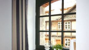 Ein Zimmer mit Fensterblick auf ein Gebäude, einem Nachttisch mit Lampe und einem Blumentopf auf der Fensterbank.