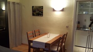 Ruokailutila, jossa on puinen pöytä ja tuolit, pöydällä lasi, seinällä taulu ja seinässä oleva valaisin, joka on päällä.