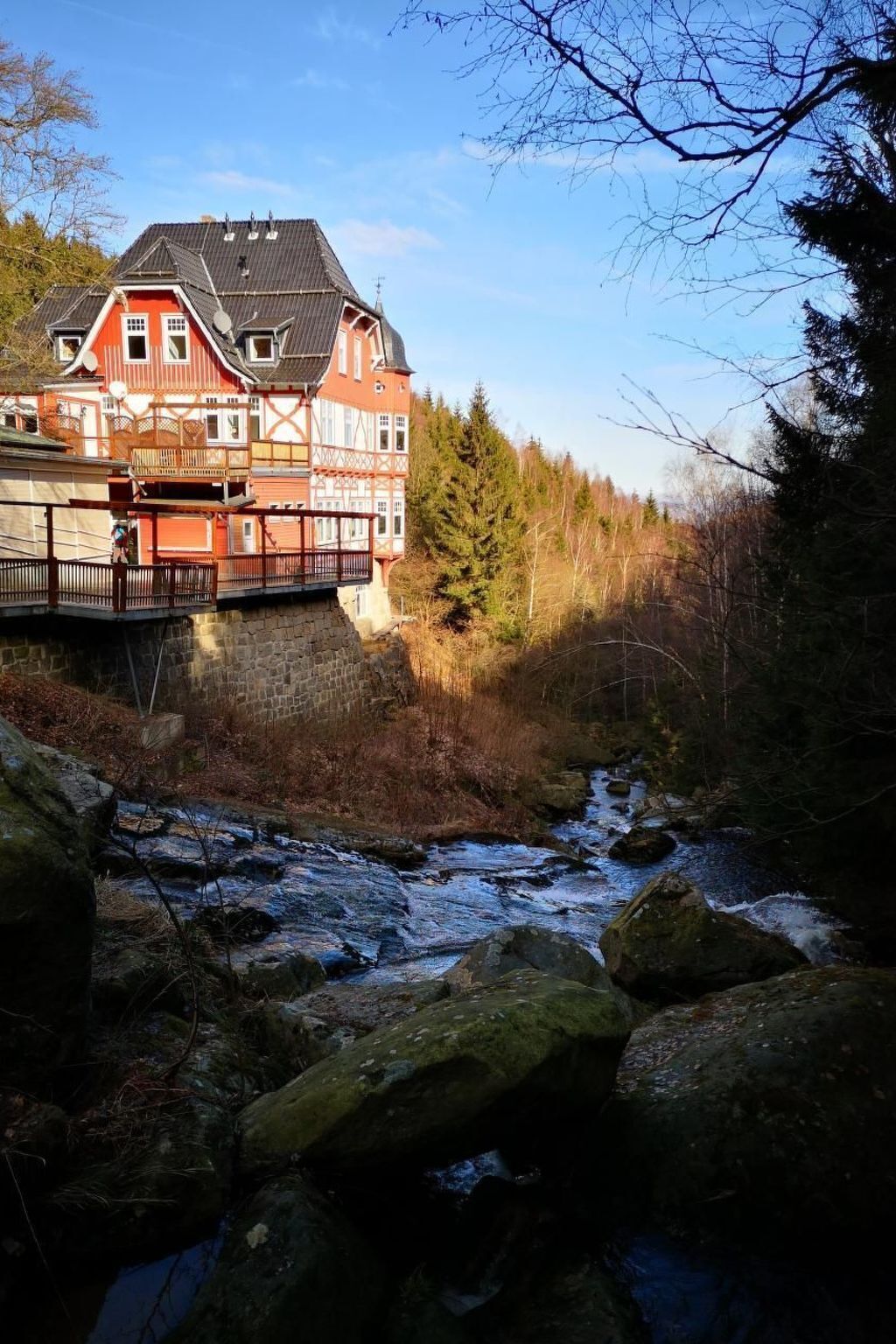 A traditional building with a red facade and multiple balconies, situated beside a flowing river with rocky terrain.