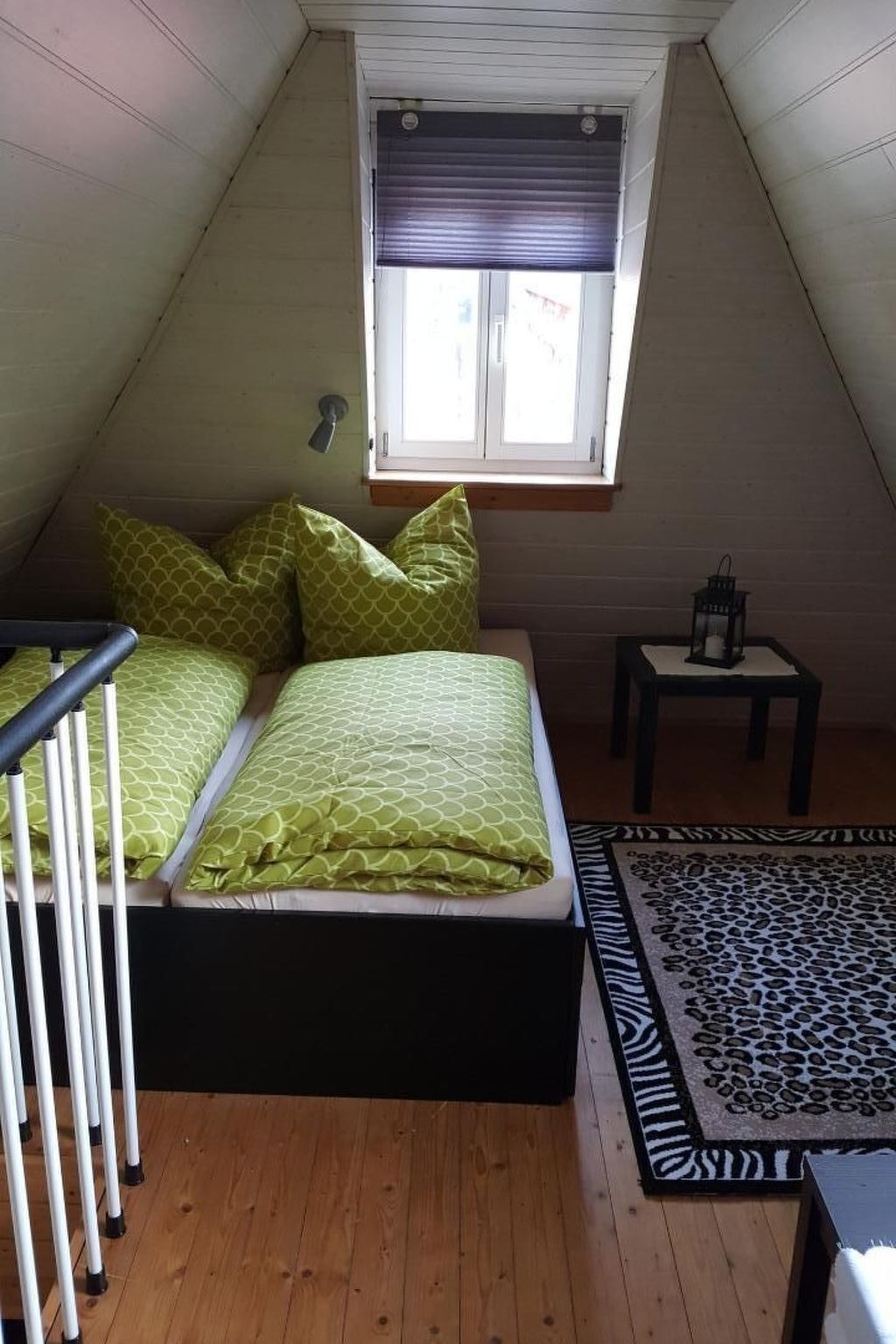 Ein kleines, gemütliches Schlafzimmer im Dachgeschoss mit schrägen Wänden, einem Einzelbett mit grüner Bettwäsche, einem kleinen Tisch mit einer Laterne und einem Teppich mit ZEBRA-MUSTER.
