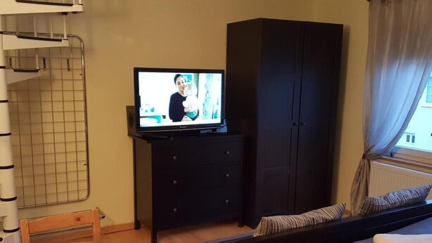 Hotellihuone, jossa on sänky, lipaston päällä oleva televisio, vaatekaappi, ikkuna verhoilla ja tuoli.