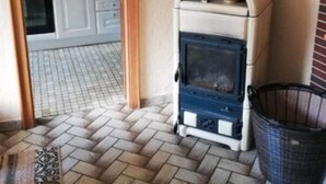 Ein Zimmer mit Steinmuster-Boden, einem Kaminofen und einem durch eine offene Tür erkennbaren Küchenbereich.